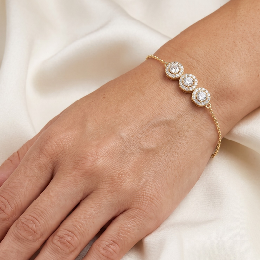 A close-up of a gold chain Moissanite bracelet with three round halo-set clear stones worn on a person's wrist, against a soft cream satin fabric background.