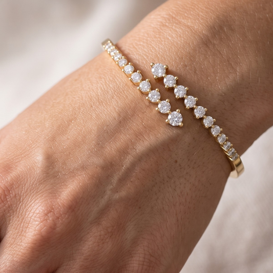 A close-up of a person's wrist wearing two delicate gold moissanite bracelets adorned with sparkling clear gemstones, set against a softly blurred light background.