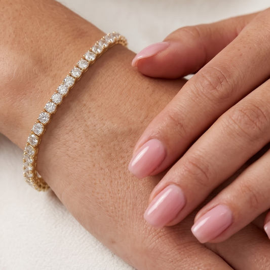 A close-up of a delicate gold tennis bracelet adorned with clear pavé-set round moissanite diamonds, shown worn on a hand with light pink manicured nails.