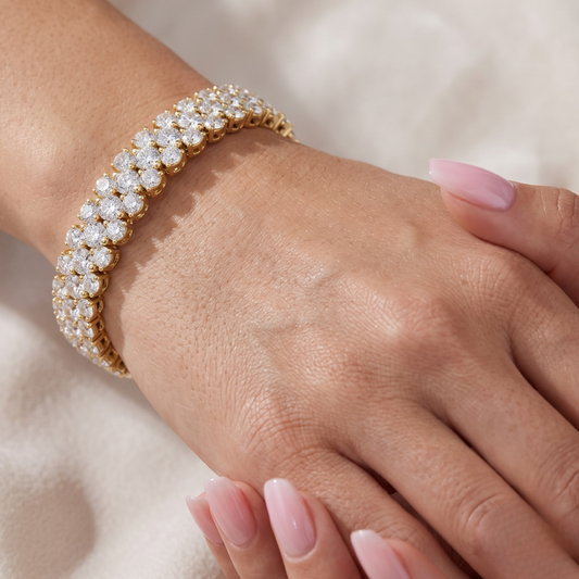 A close-up of a gold tennis bracelet adorned with sparkling clear moissanite gemstones, worn on a woman's wrist with her hand resting nearby against a soft, light beige background.
