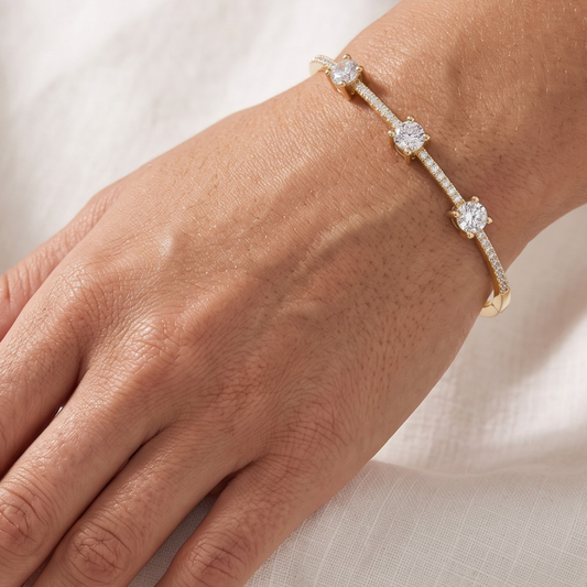 A delicate gold bracelet with three round-cut clear moissanite gemstones and pavé accents, shown worn on a person's wrist against a soft white fabric background.