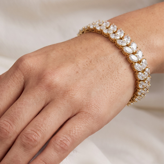 A close-up of a gold tennis bracelet with clear moissanite gemstones worn on a person's wrist, set against a soft white fabric background.