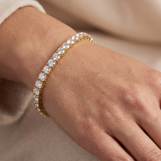 A close-up of a gold tennis bracelet with clear round-cut moissanite stones worn on a person's wrist, set against a soft beige background.