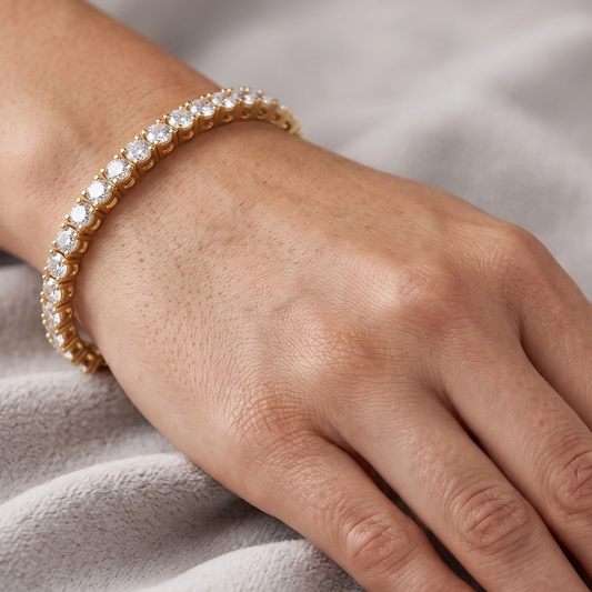 A close-up of a gold tennis bracelet with clear round-cut moissanite stones worn on a person's wrist, set against a soft gray fabric background.