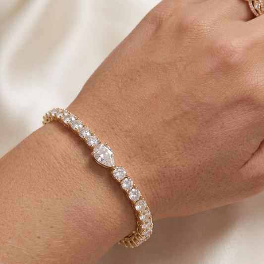 A gold tennis bracelet with clear moissanite gemstones worn on a person's wrist, shown against a soft cream-colored fabric background.