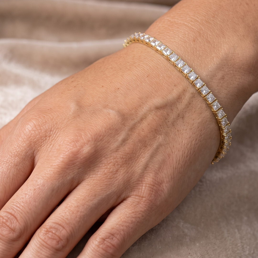 A close-up of a gold tennis bracelet with square-cut clear moissanite  stones worn on a person's wrist, set against a soft beige fabric background.