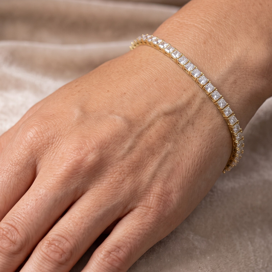 A close-up of a gold tennis bracelet with square-cut clear moissanite  stones worn on a person's wrist, set against a soft beige fabric background.