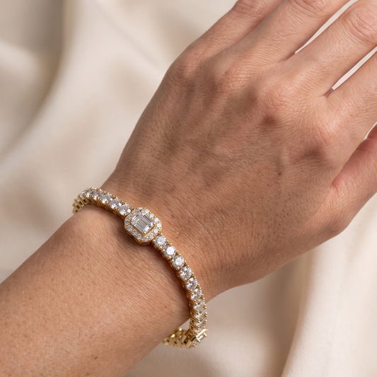 A close-up of a gold tennis bracelet with a central cushion-cut moissanite diamond and surrounding small diamonds, worn on a person's wrist against a soft cream fabric background.