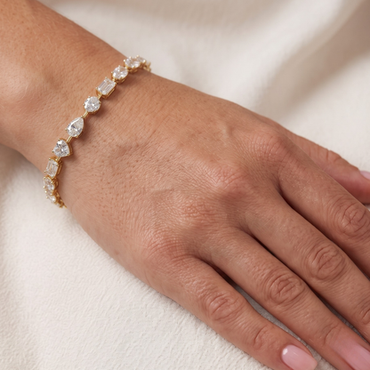 A gold bracelet with clear teardrop-shaped moissanite diamond worn on a person's wrist, shown against a soft cream-colored fabric background.