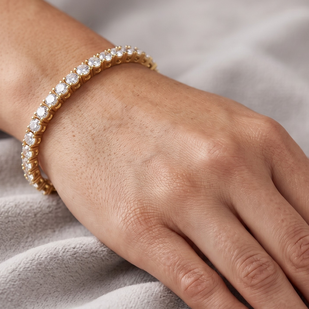 A close-up of a gold tennis bracelet with clear round-cut moissanite stones worn on a person's wrist, set against a soft gray fabric background.